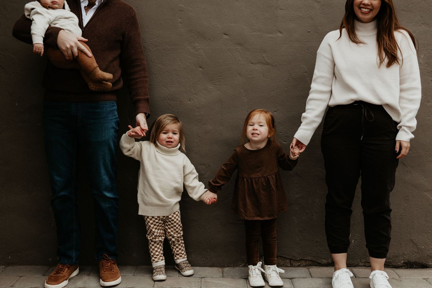 Family of four holding hands against a gray wall, parents with two toddlers in coordinating neutral outfits. Miranda Photo & Co