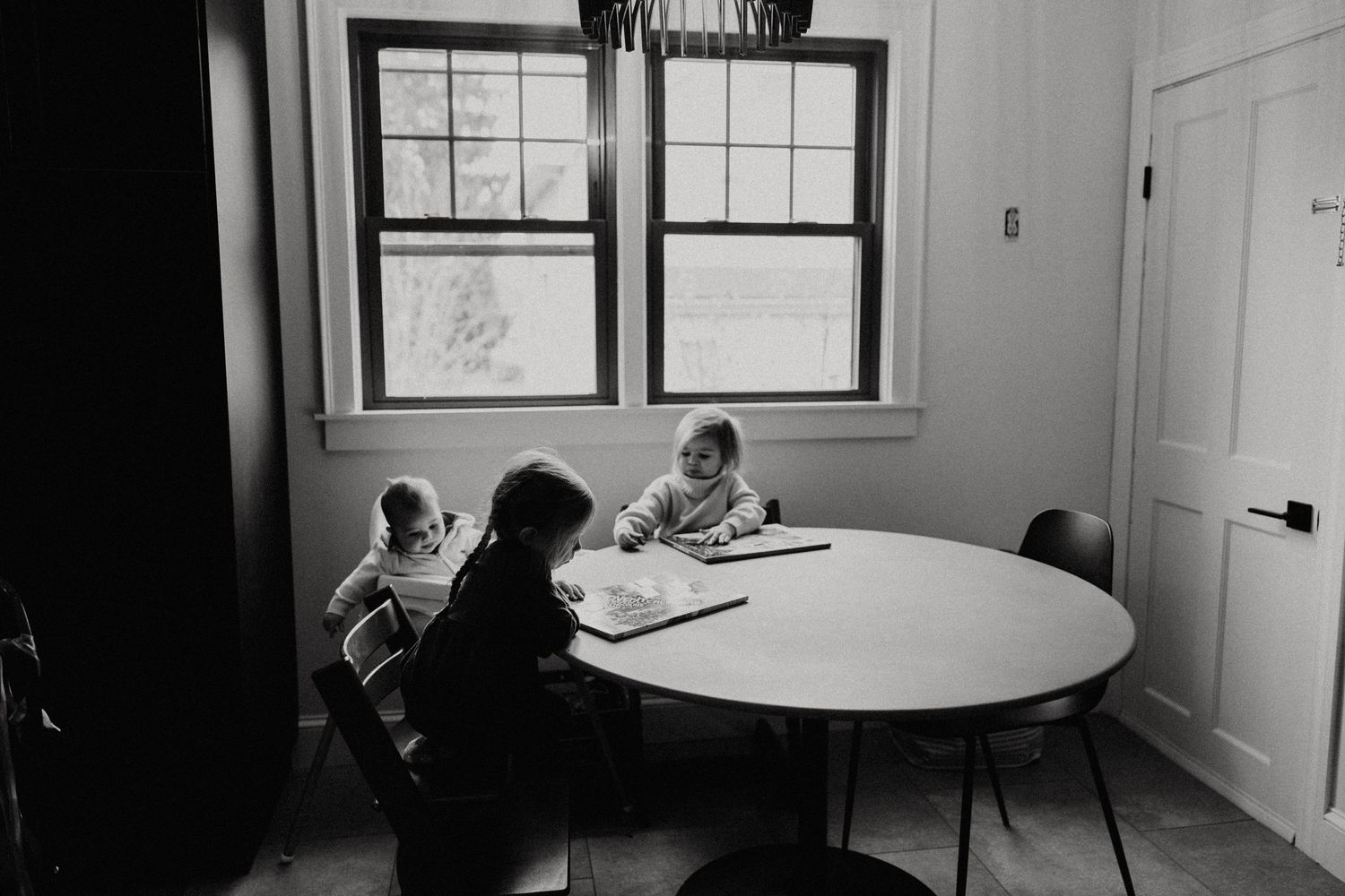 Black and white photo of three children sitting at a round table near windows, drawing or doing homework together. Miranda Photo & Co