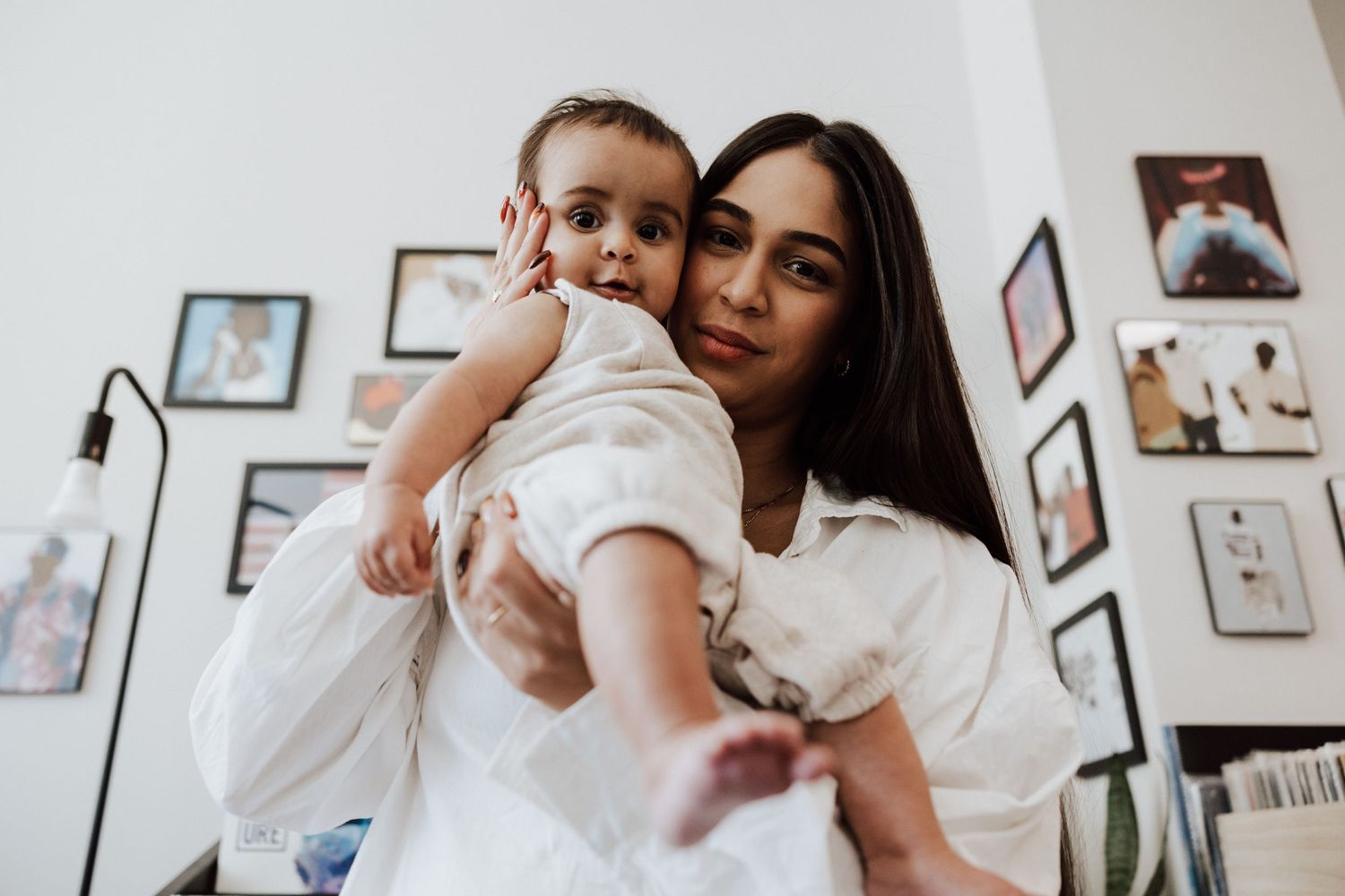A young mother with long dark hair holds her baby in a white room with framed photos on the wall. Miranda Photo & Co