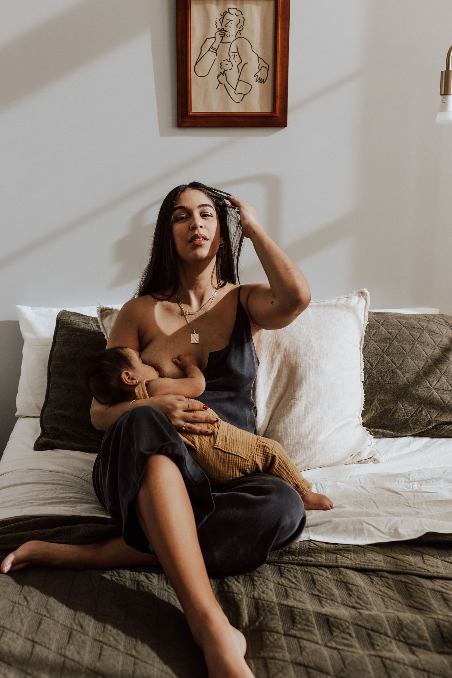 A woman in a dark slip dress sits on a bed breastfeeding her baby, with artwork on the wall behind her. Miranda Photo & Co