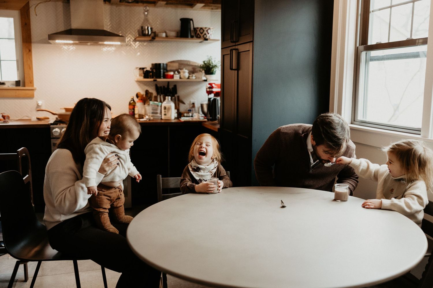 A joyful family laughs together around a round kitchen table in a cozy home setting. Miranda Photo & Co