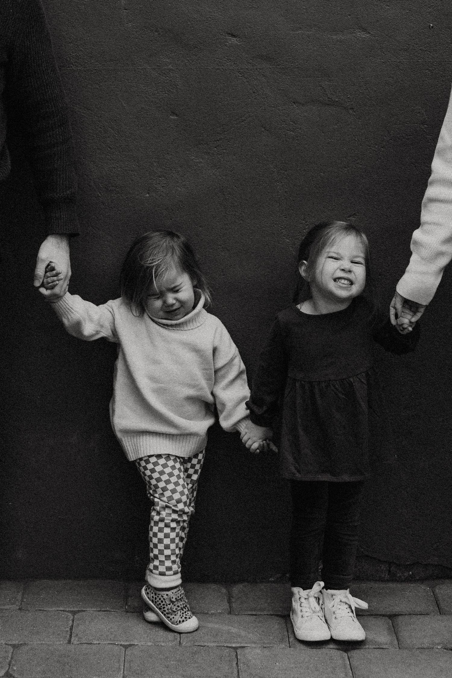 Black and white photo of two young sisters holding hands and smiling against a dark wall. Miranda Photo & Co
