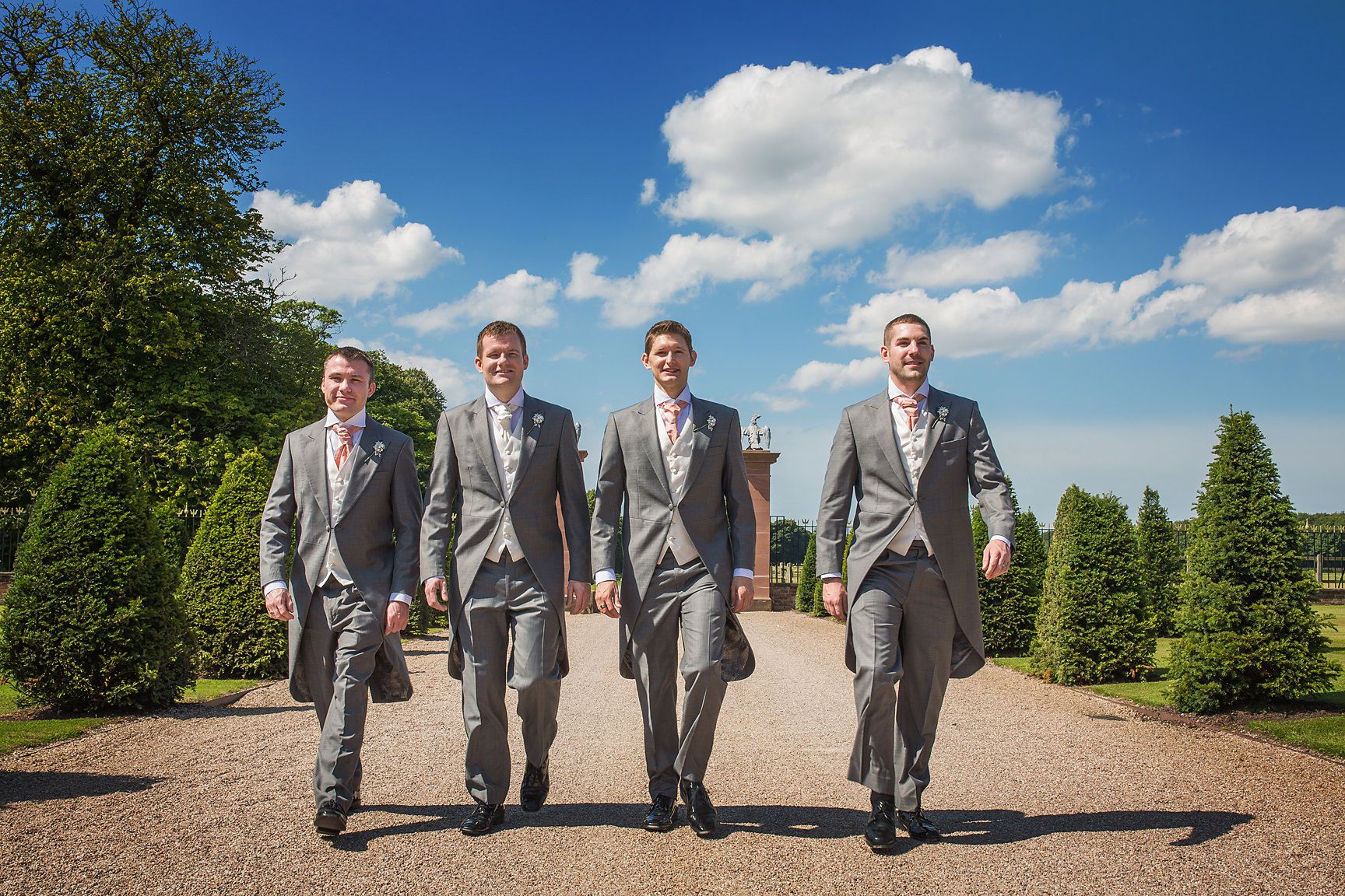Four groomsmen in matching gray suits walk together on a path under blue skies.