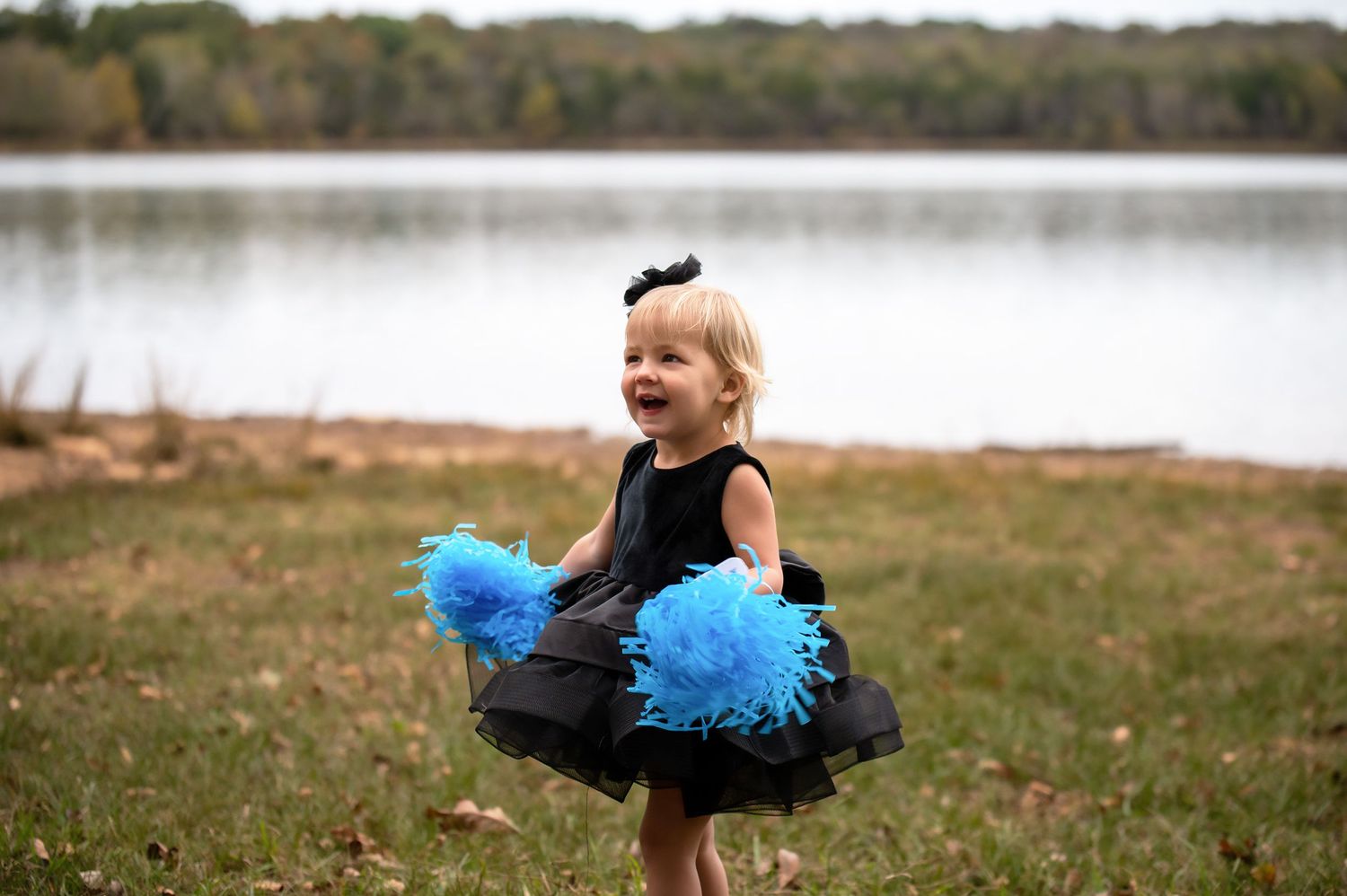 A small dancer in a black tutu twirls with blue pom poms by a serene lake.