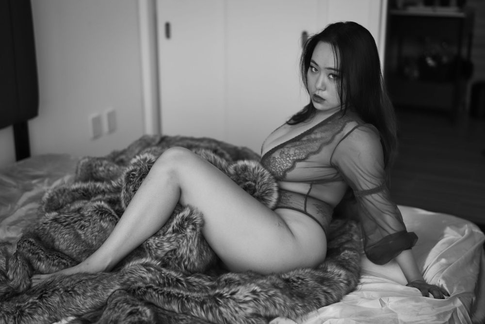Artistic black and white photograph of a person in sheer lingerie posing on textured bedding.