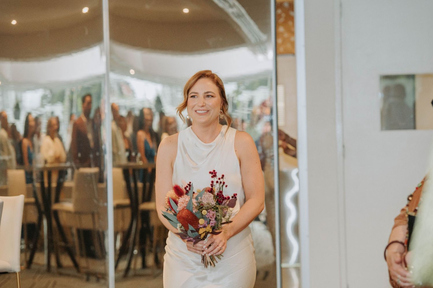 A bride in a sleeveless white dress holds a red floral bouquet while walking.