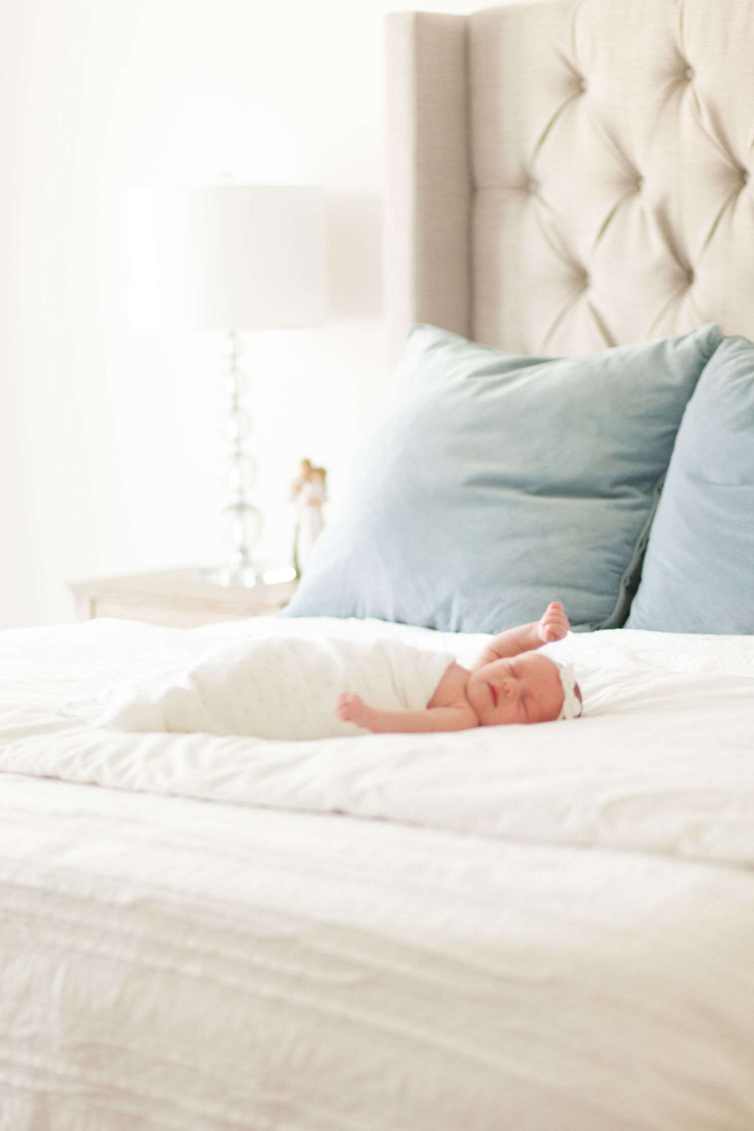 A baby lays in a cozy bedroom with a tufted headboard, white bedding, and soft blue accent pillows in natural light.