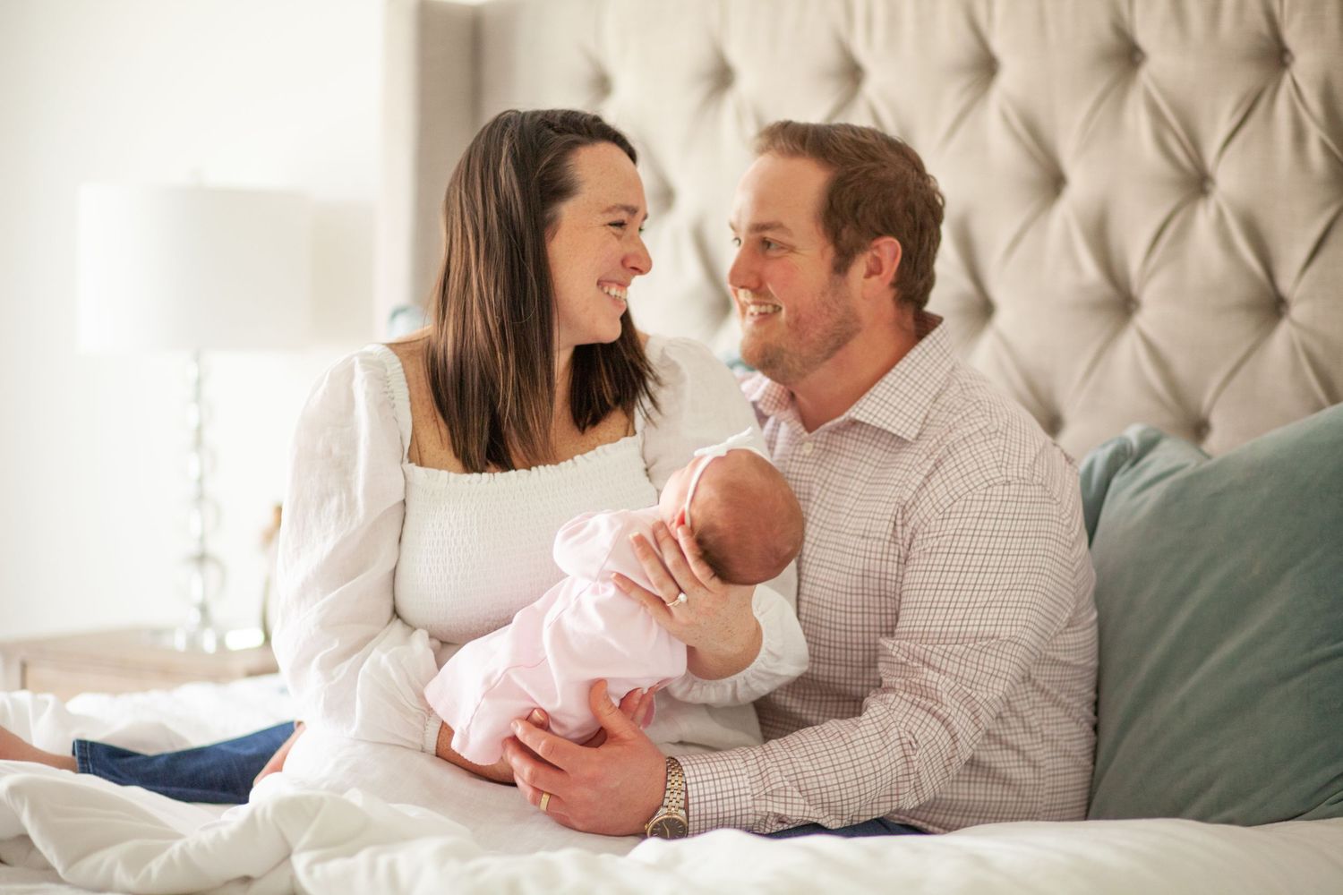 A couple sits on their bed sharing a tender moment with their newborn baby dressed in pink at their Raleigh NC family session.