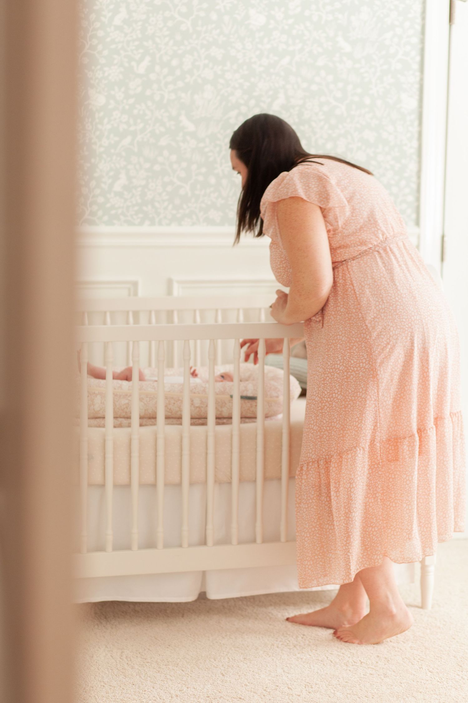 A person wearing a pink dress stands next to a white crib in a nursery with soft lighting and cream walls at their Raleigh newborn photos.
