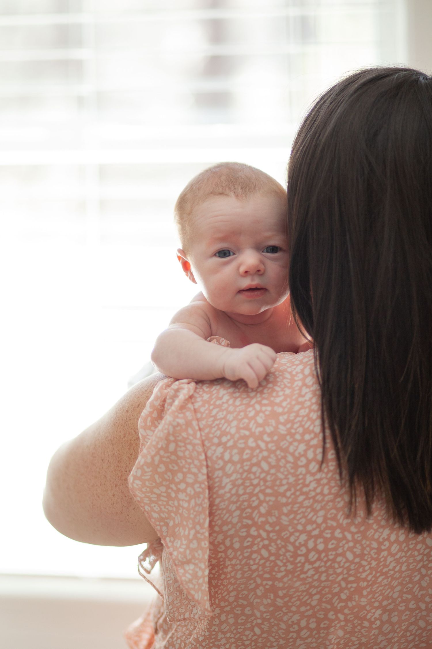 Baby peers over shoulder of parent wearing peach floral dress in soft window light setting.