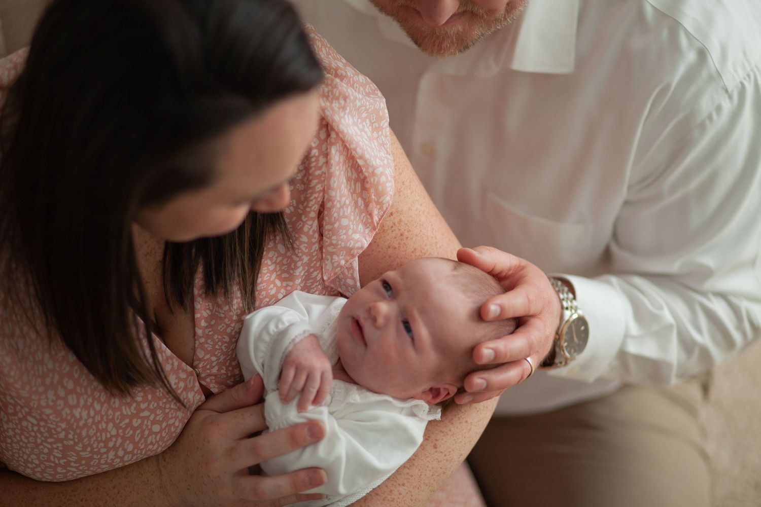 A tender moment between parents and their newborn baby swaddled in white, with soft lighting and warm tones.