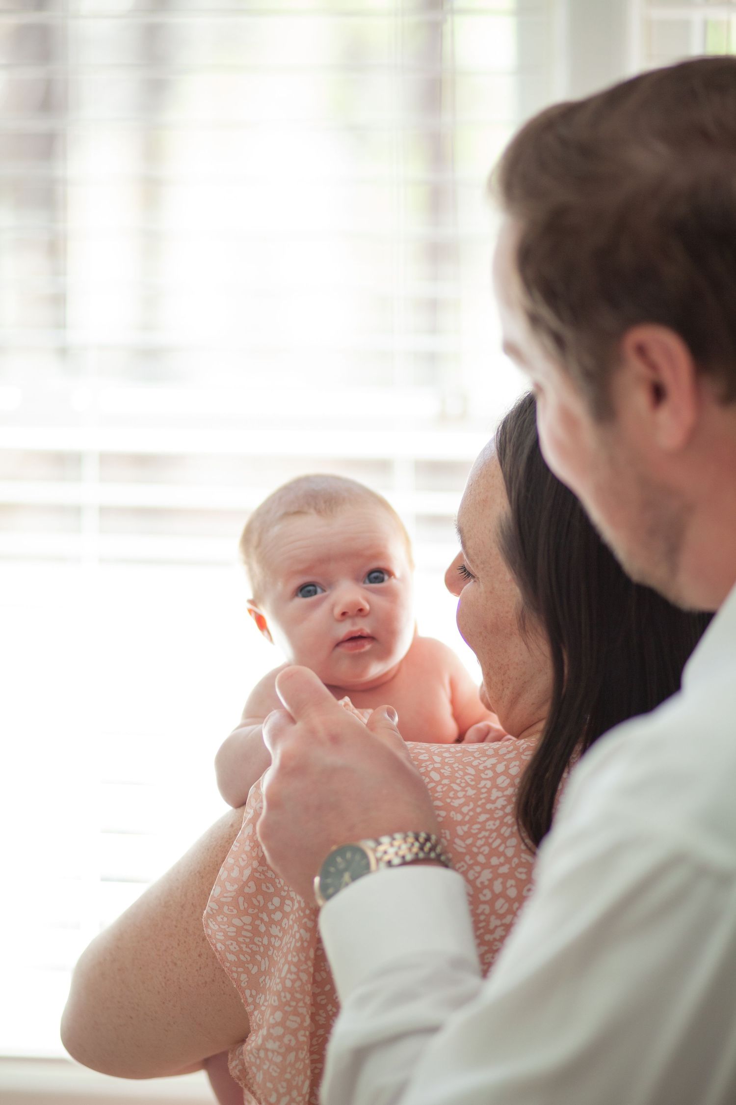 Baby wearing pink dress gazes up while being held against sunlit window at her Raleigh at home newborn session