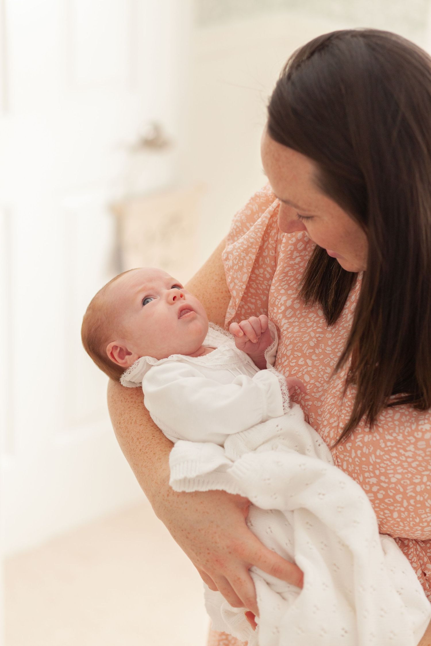 A tender moment between a mother in a peach dress and her newborn baby wearing white, gazing at each other lovingly.