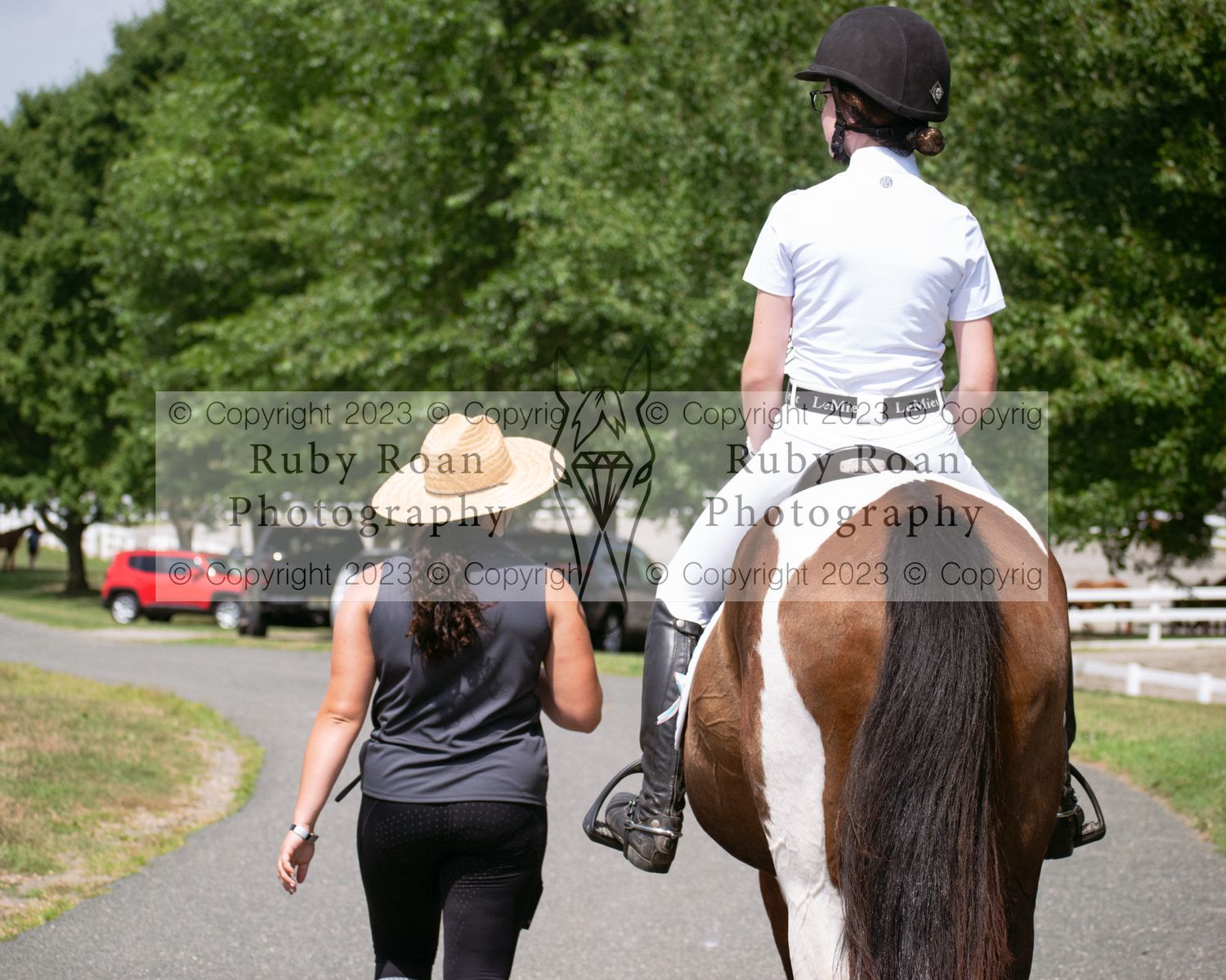 woman in hat walking to the left of girl in white on brown and white horse