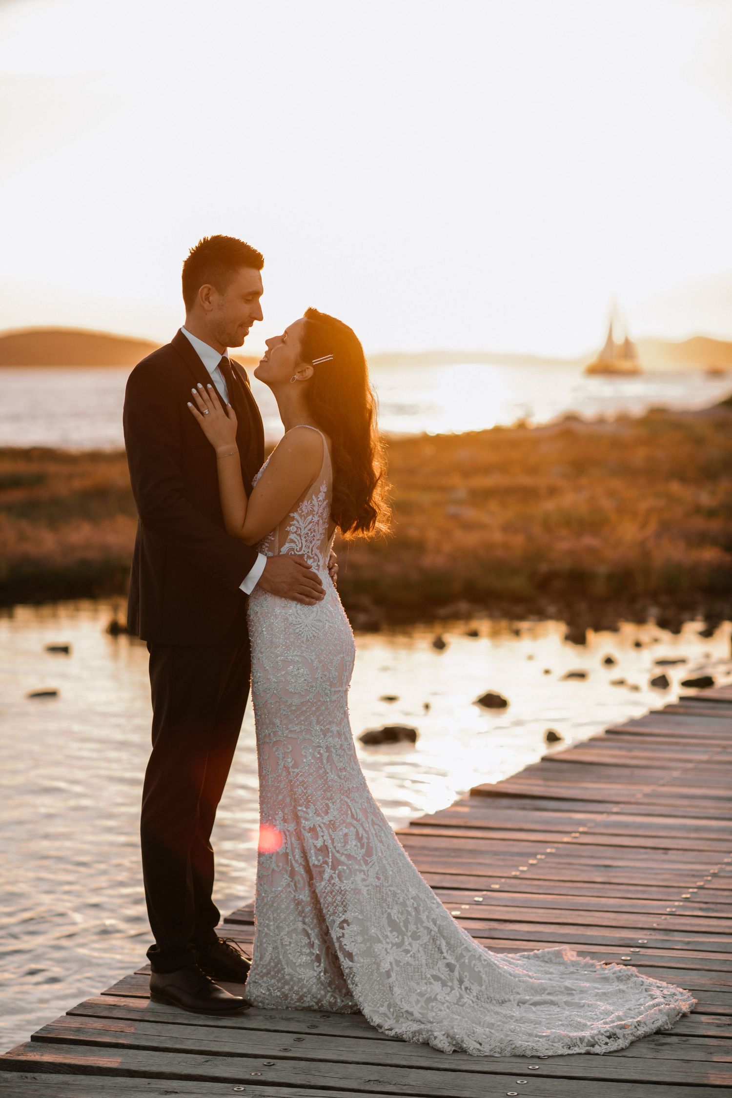 Newlywed couple embraces on wooden dock at sunset with ocean and golden light in background.