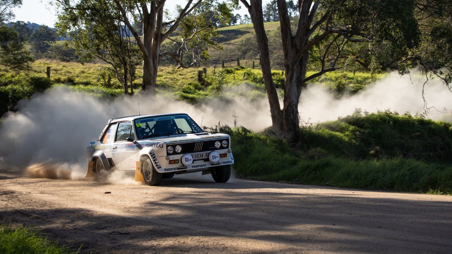 Classic white rally car kicking up dust while drifting on a dirt road.