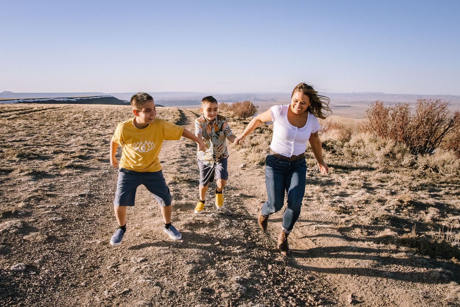 Three people jumping joyfully on desert trail with mountains in background.