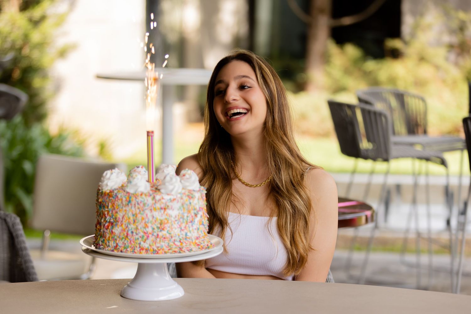 Smiling young woman celebrating birthday with sparkler cake at outdoor patio table.