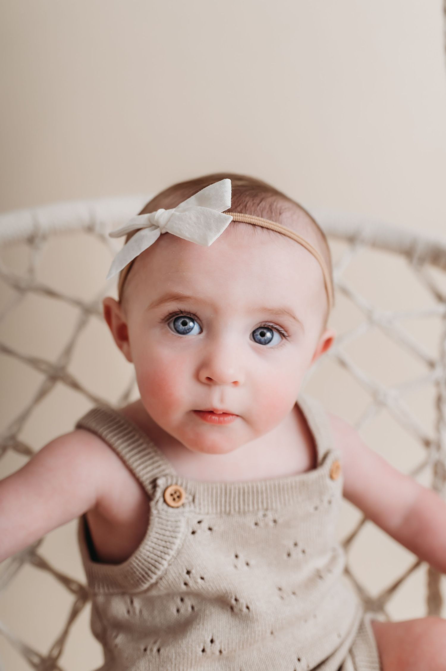 9-month-old girl with big blue eyes looks up sweetly from macrame swing in a white bow and knit romper.