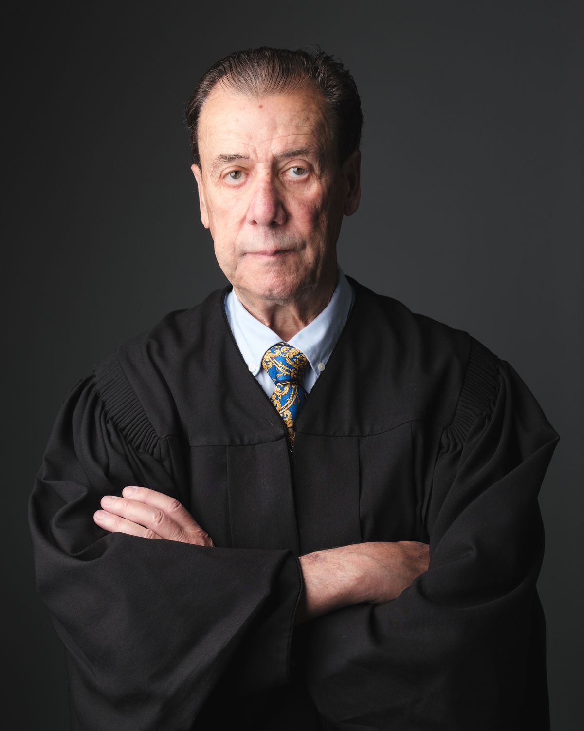 Professional portrait of a judge in traditional black robes with a blue and gold tie against a dark background.