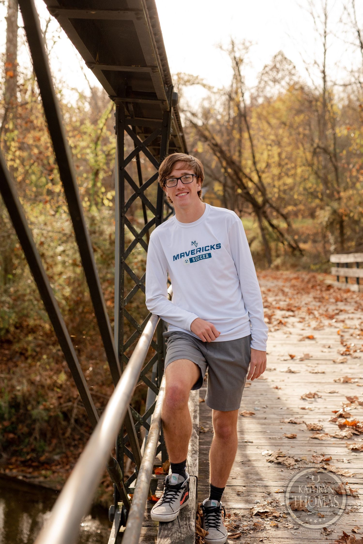 A person poses on an old metal bridge with autumn trees in the background.