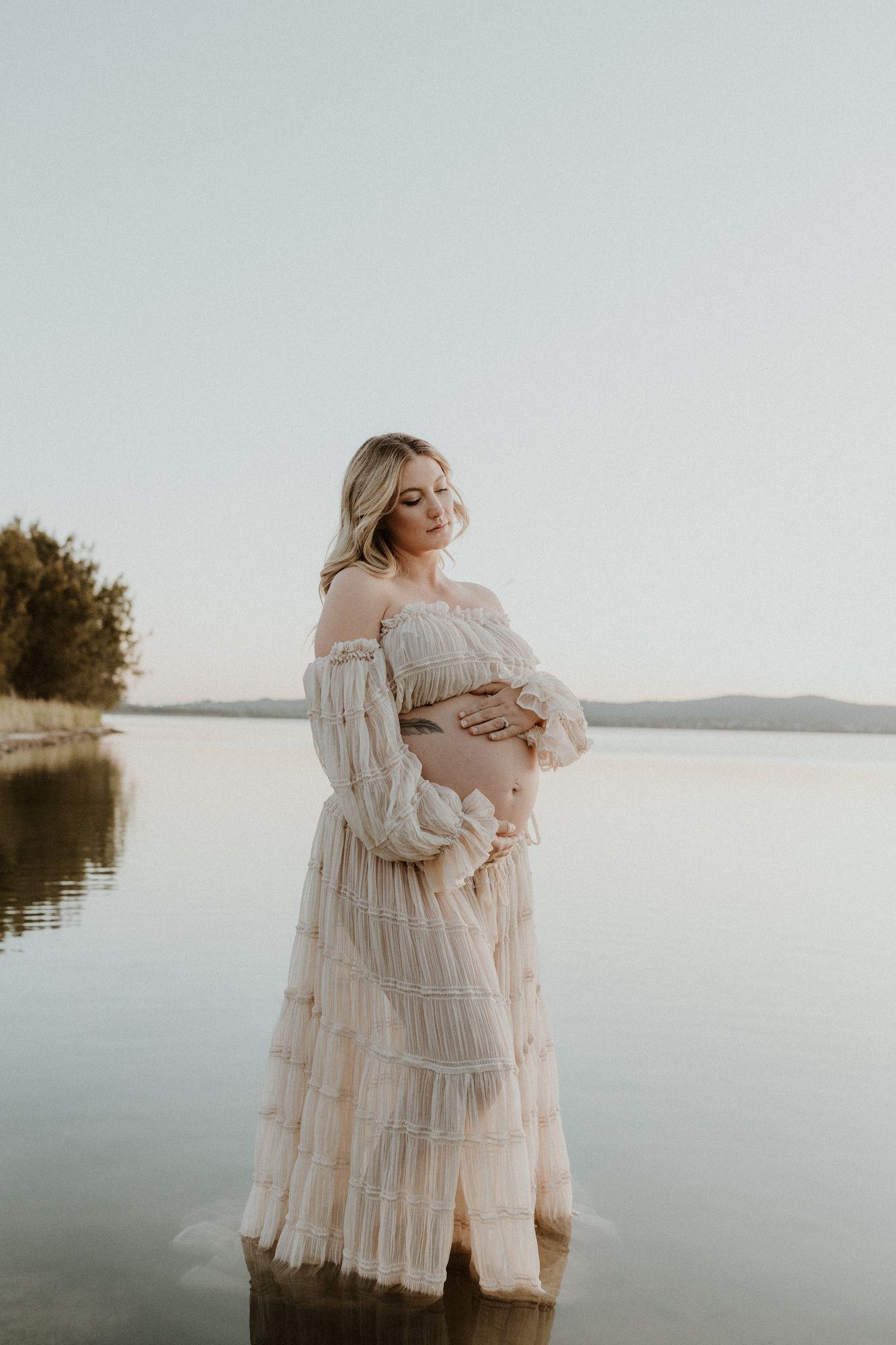 Ethereal maternity portrait of an expectant mother in a flowing white dress standing in calm waters.