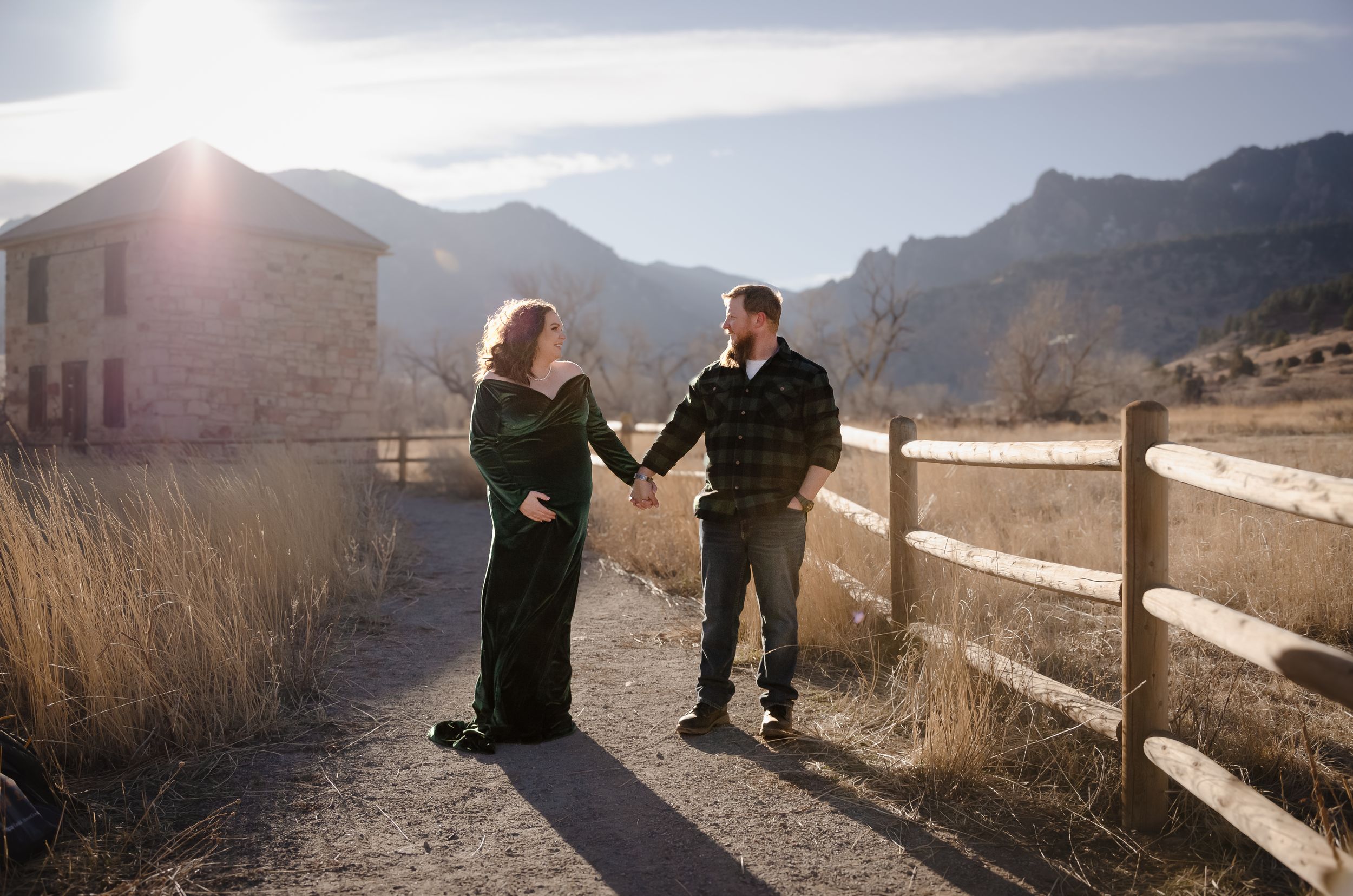 Couple sharing intimate moment on mountain path with rustic fence and sunlight.