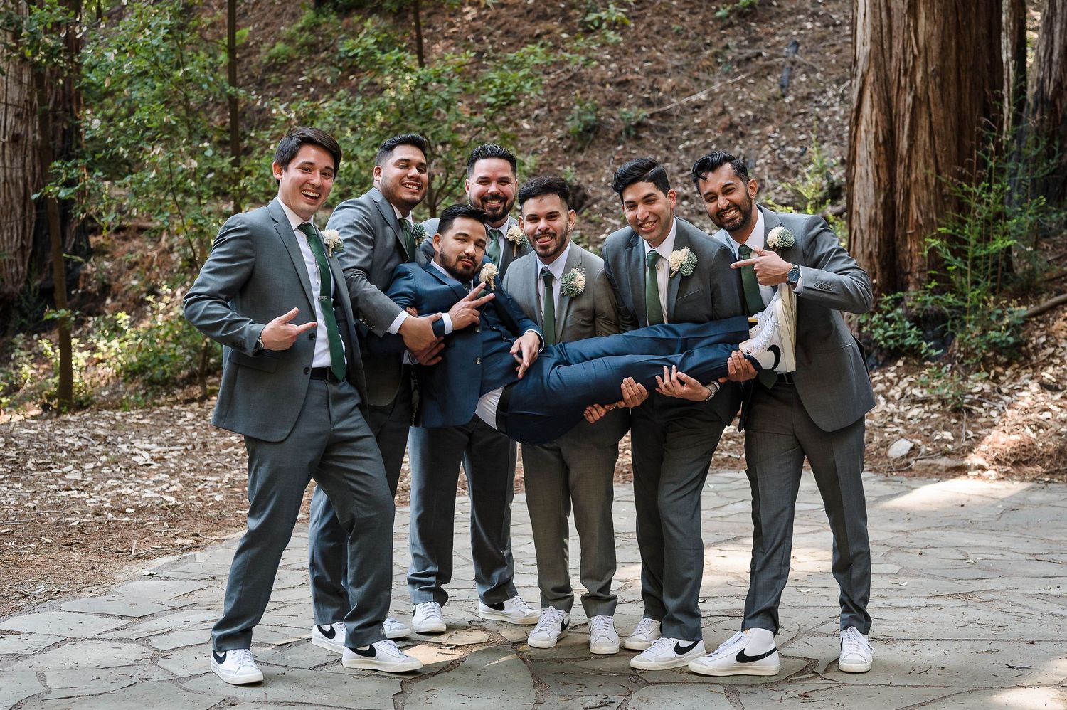 A cheerful wedding party in grey suits poses playfully on a forest path.