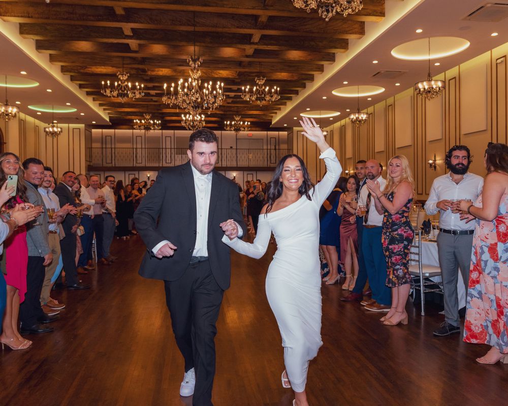 Newlyweds make their grand exit through a crowd of guests in an elegant ballroom with chandeliers and wood flooring.