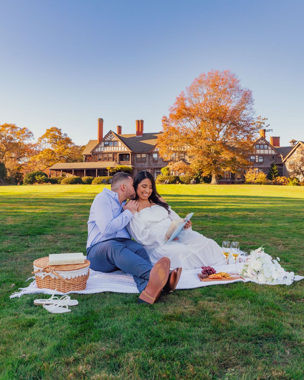 A couple enjoys a romantic fall picnic on a blanket with snacks on a lawn in front of a grand estate house.