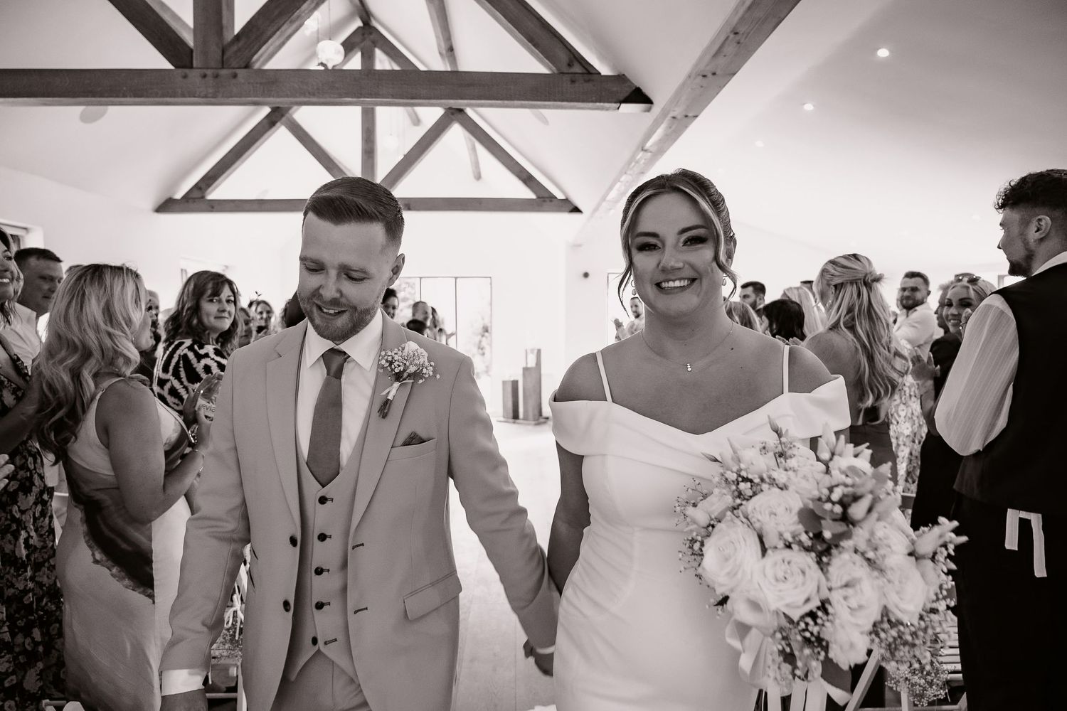 Newlyweds walk hand in hand during wedding ceremony in rustic venue with exposed wooden beams and happy guests.