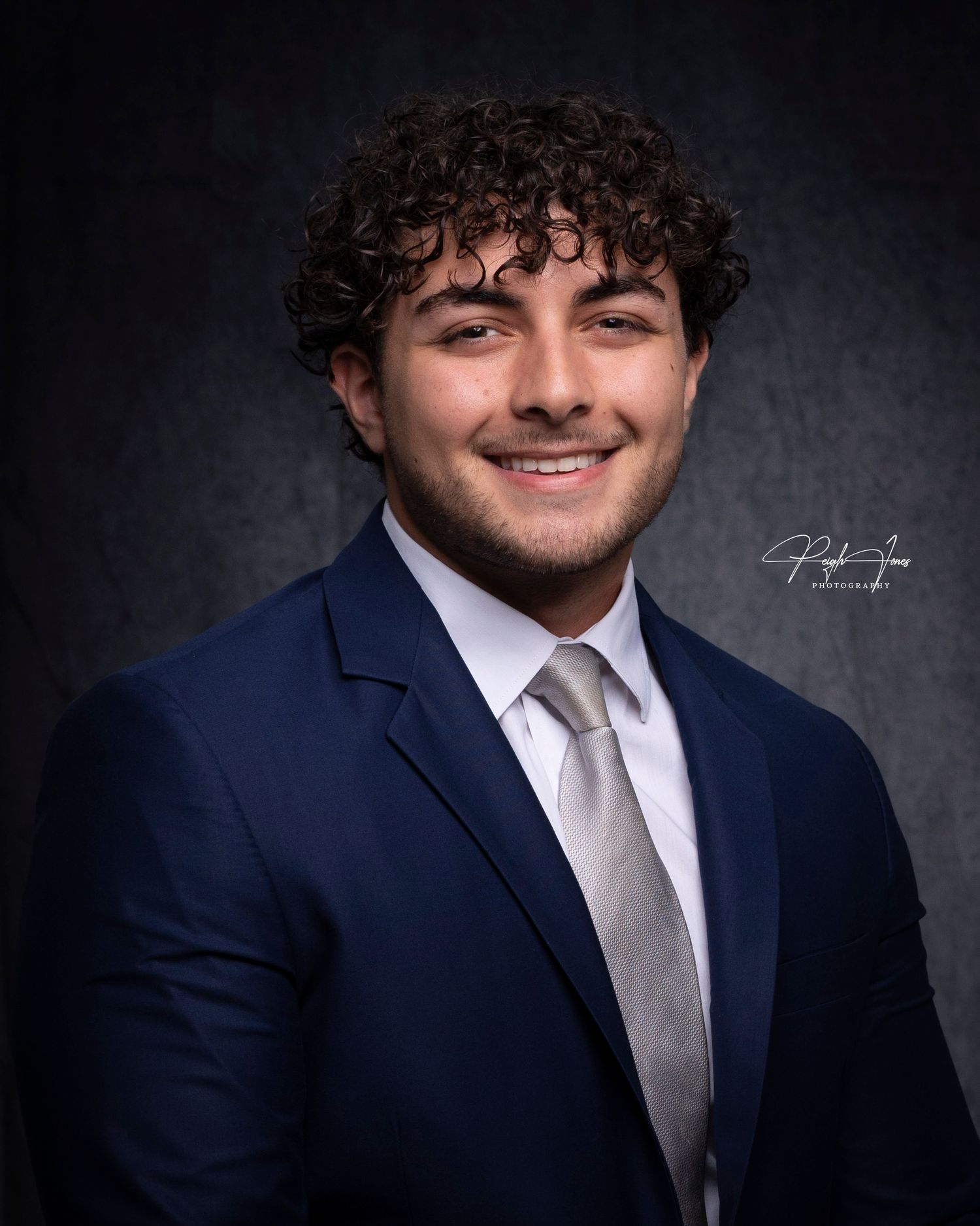 Young man with curly hair smiles in navy suit and silver tie for formal portrait.