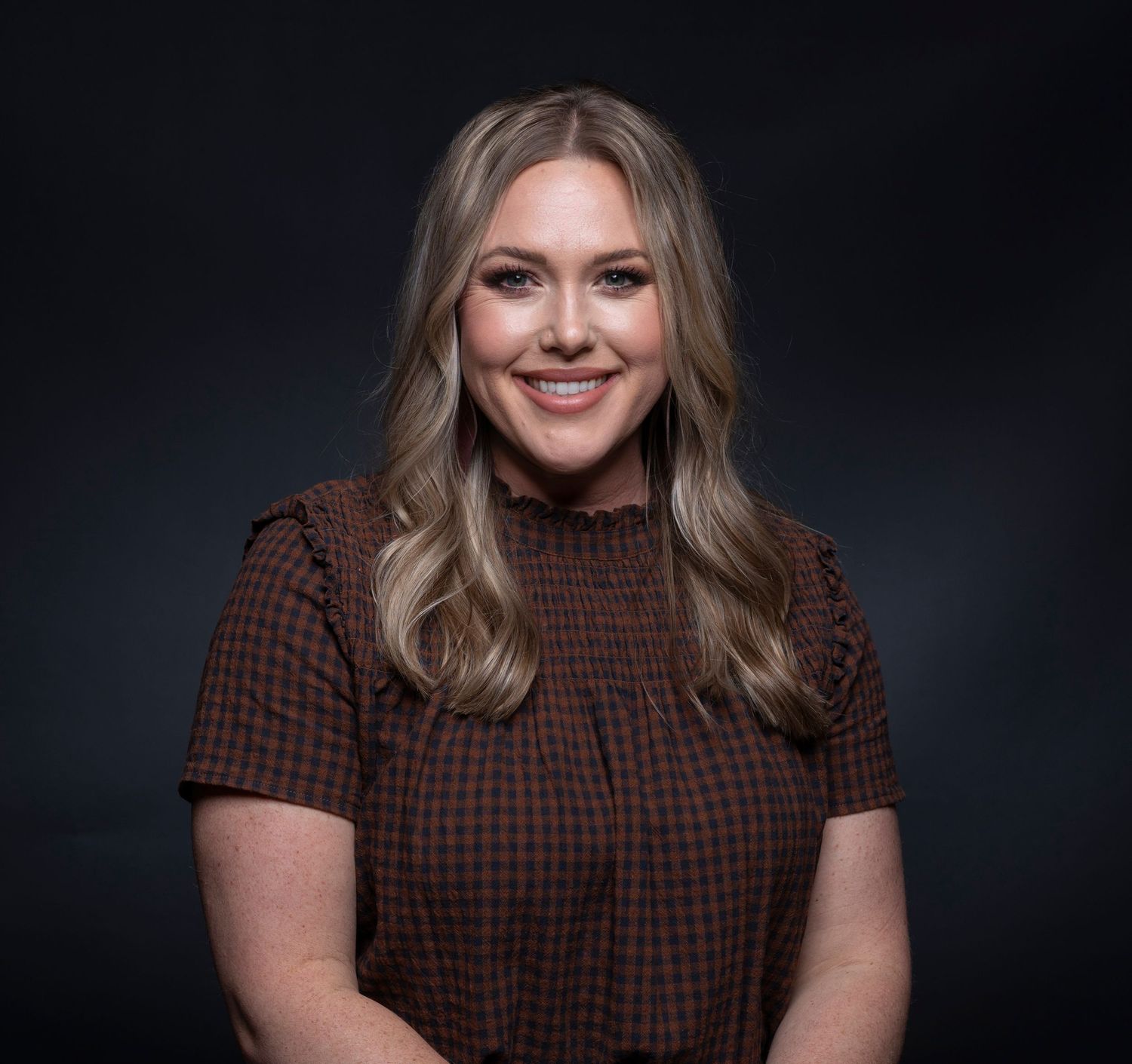 Smiling blonde woman in brown plaid top poses for professional headshot against dark background.