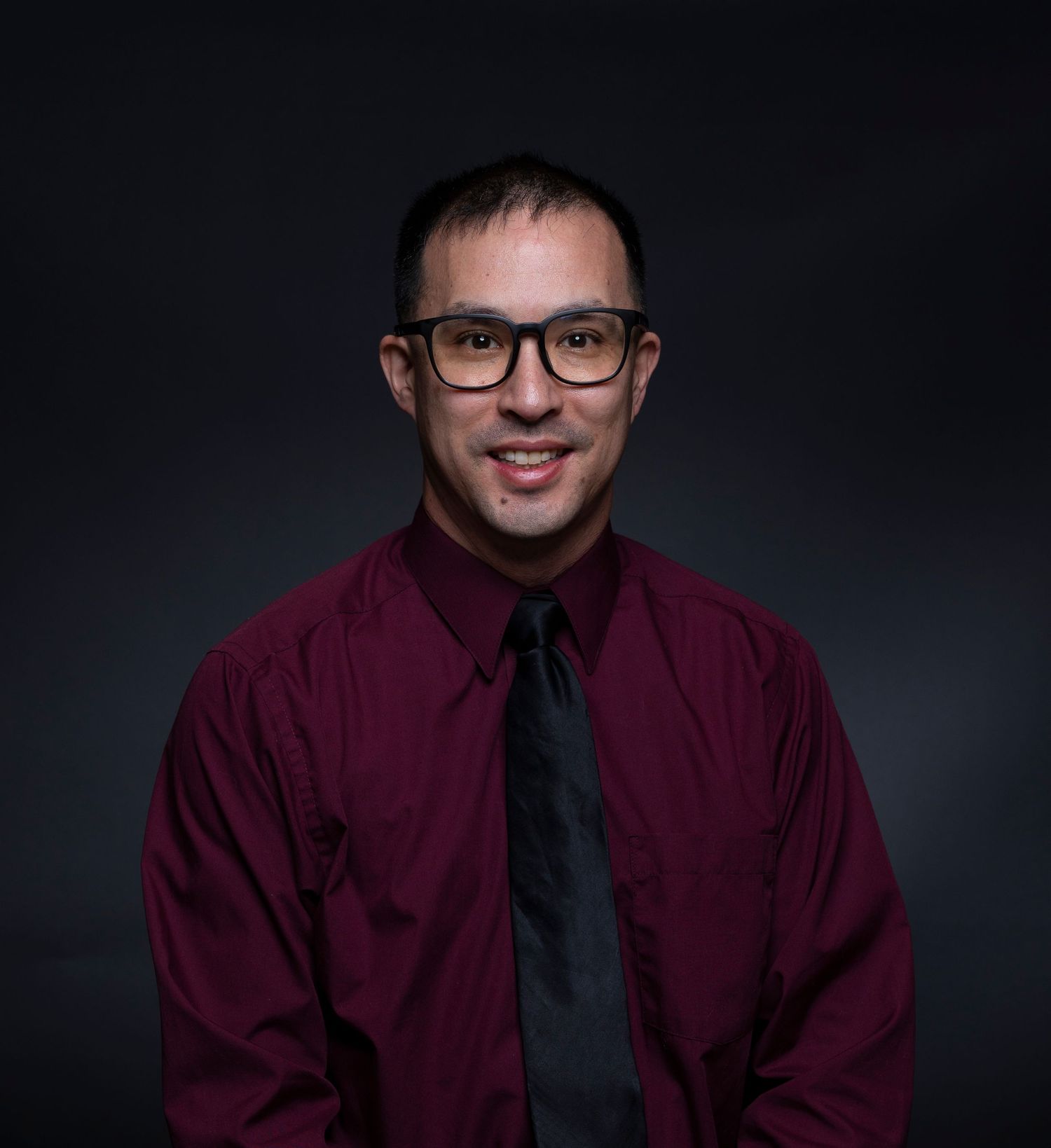 Professional man in burgundy shirt and tie smiles for a portrait against a dark background.