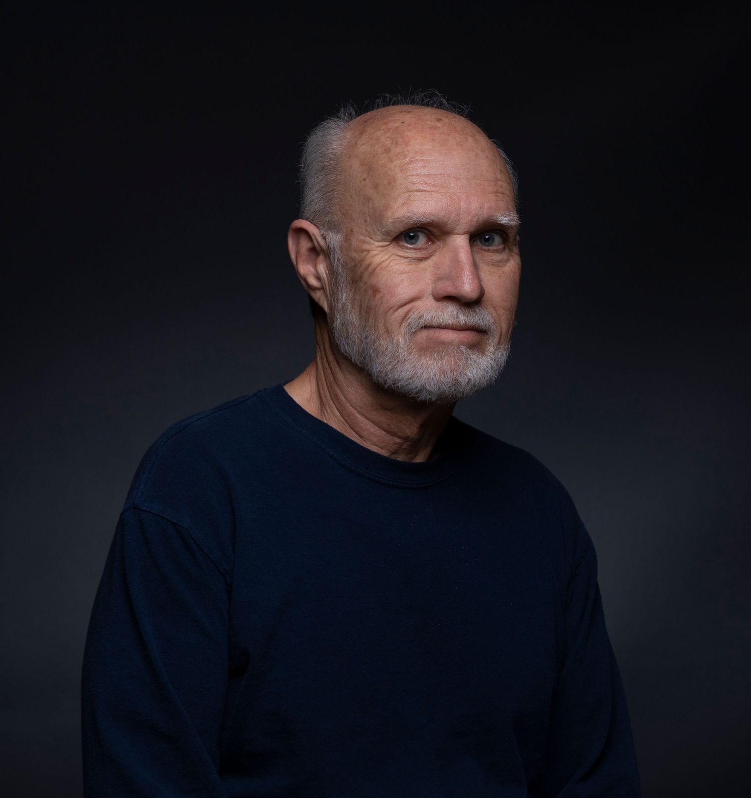 Older man with gray beard wearing navy blue sweater poses for portrait against dark background.