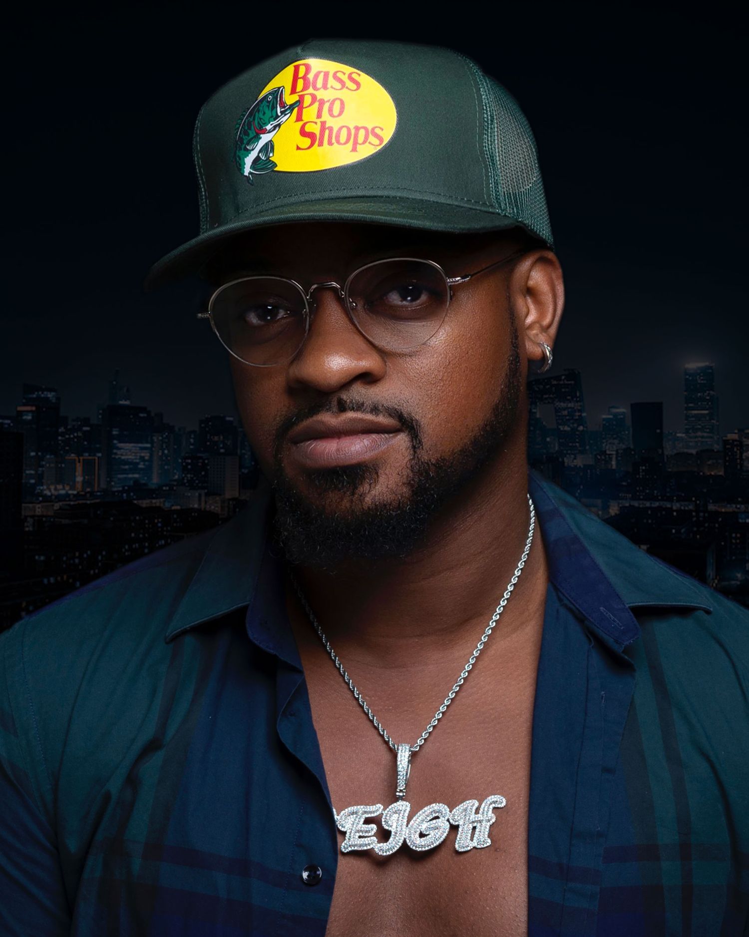 Man wearing Bass Pro Shops cap and EJOH chain necklace poses for portrait against city backdrop.