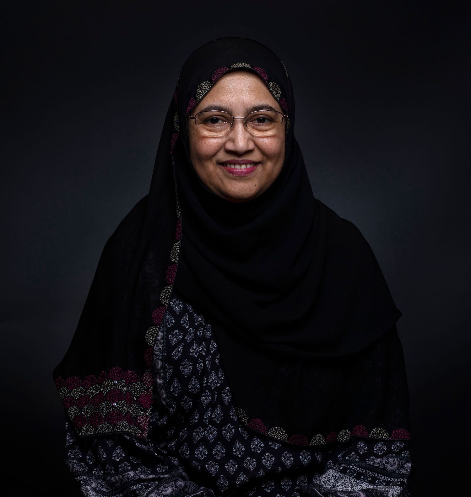 Smiling woman wearing black hijab and glasses poses for portrait against dark background.