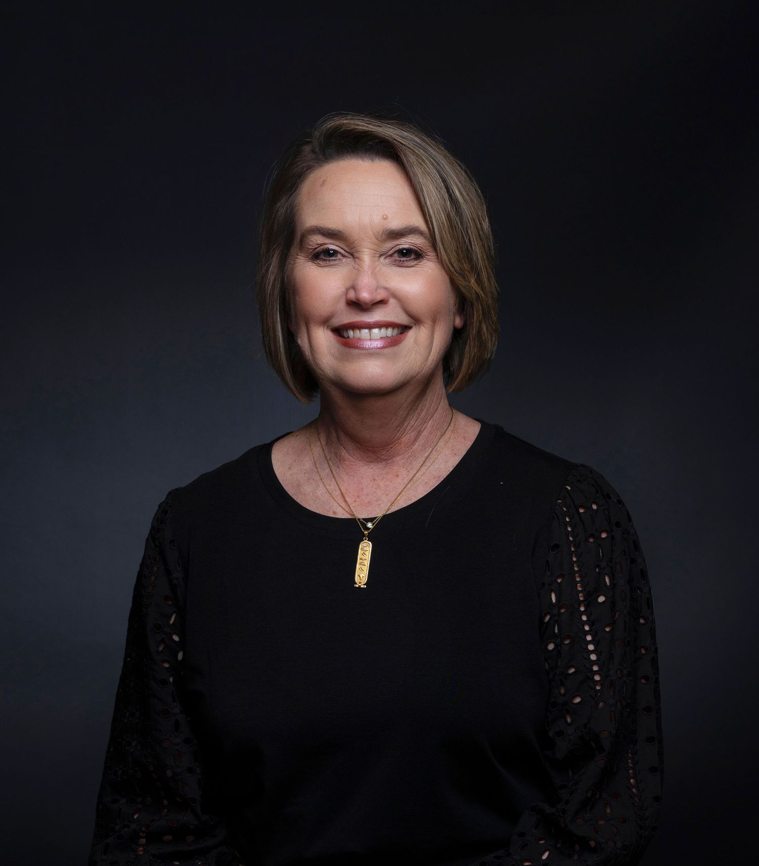 Smiling woman with short brown hair wearing black top and gold necklace poses for headshot.