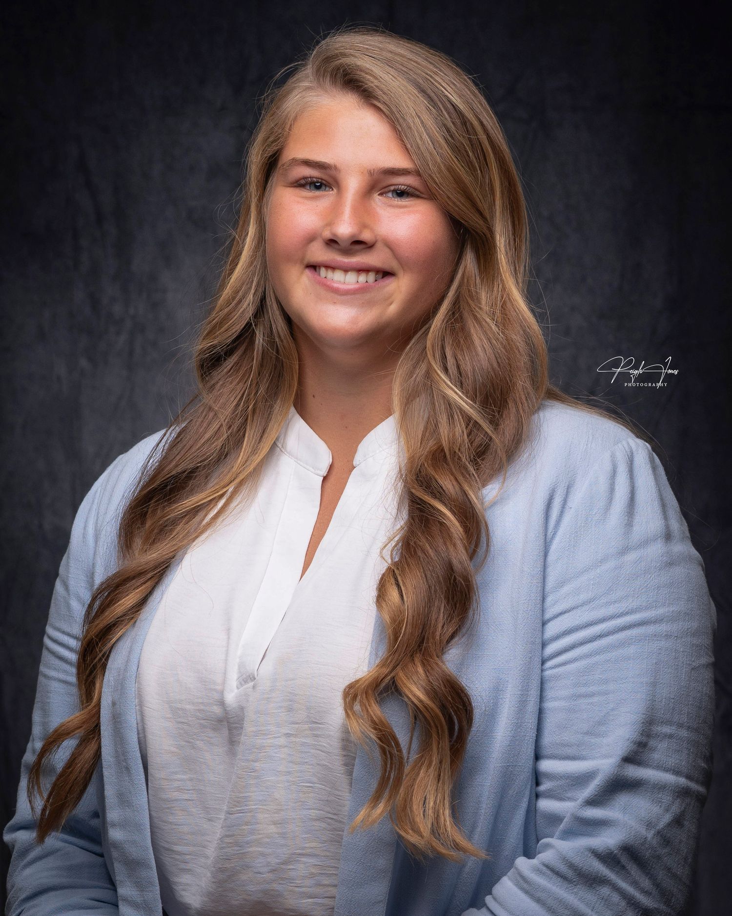 Young woman with long wavy blonde hair smiling in a light blue cardigan against a dark studio background.
