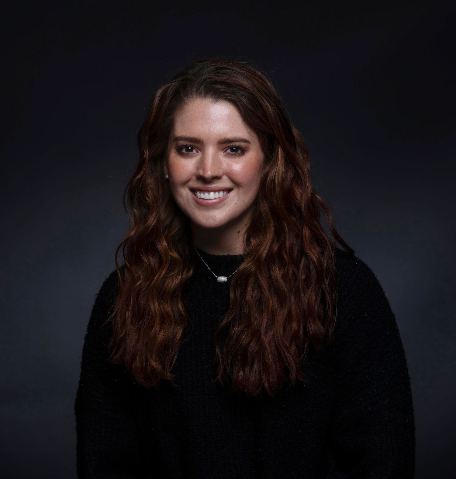 Young woman with long red wavy hair smiles for professional headshot against dark background.