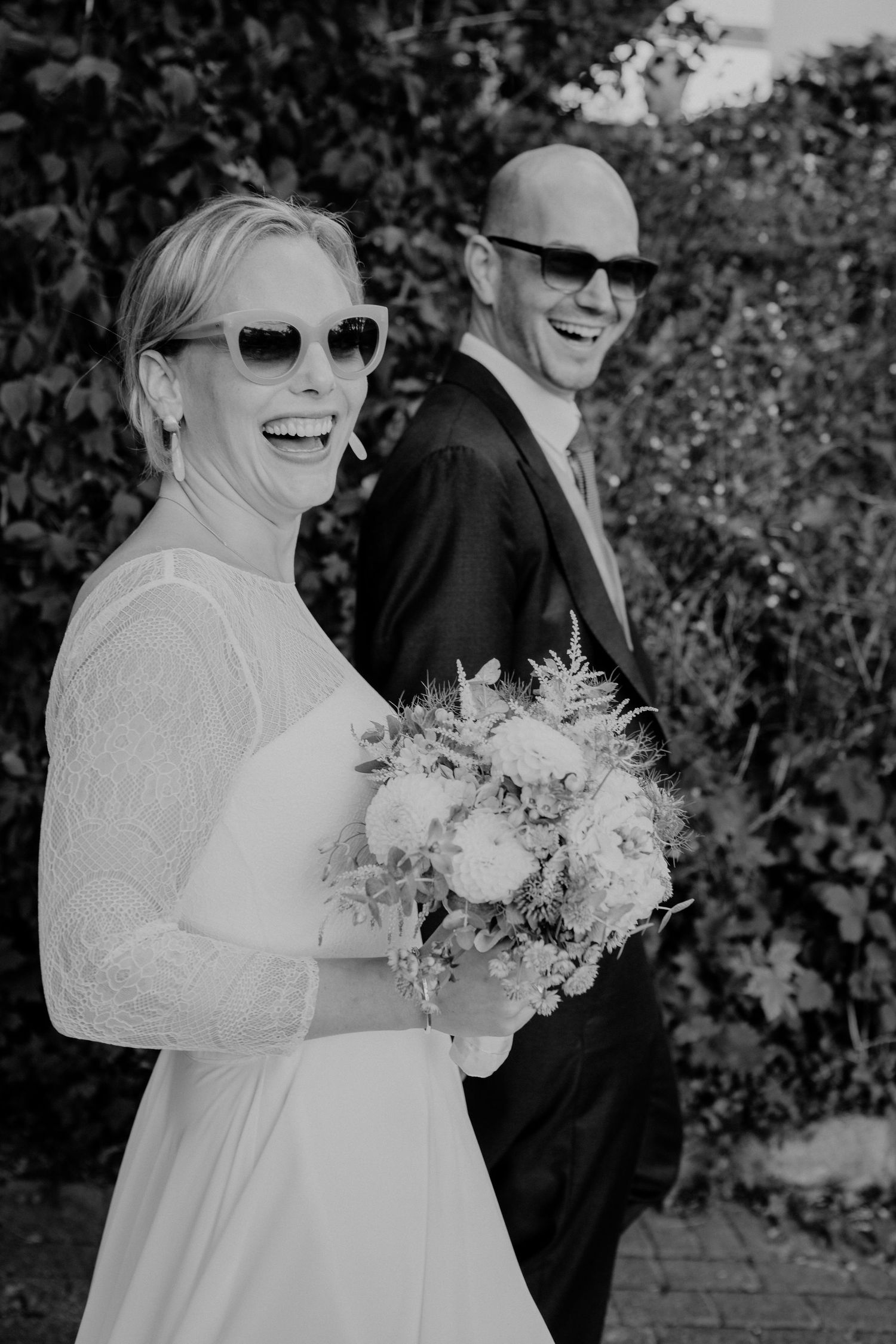 A joyful wedding couple wearing sunglasses laughs together outdoors, with the bride holding a beautiful floral bouquet.