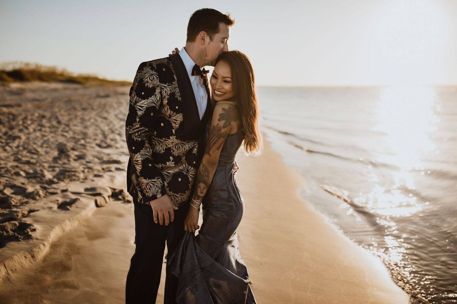 A couple embracing on a beach at sunset wearing formal attire.