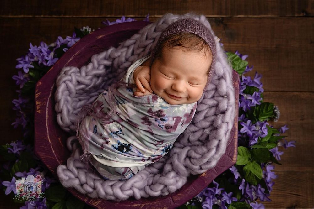 Sleeping newborn wrapped in grey knit blanket surrounded by purple flowers in wooden bowl.
