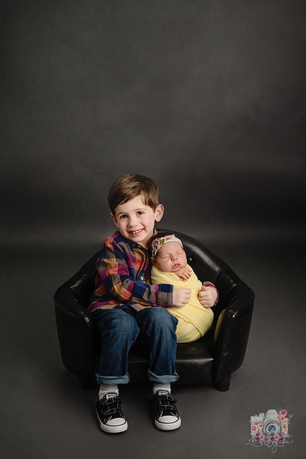Two siblings sitting together in a black leather chair against dark background.