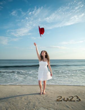 A girl in a white dress throwing her graduation cap in the sky on a beach.