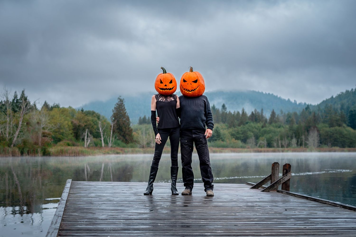 pumpkin head jack-o-lantern couple pose on a lake dock on a cloudy, moody day