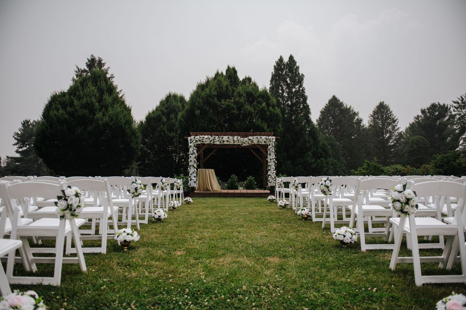 White chairs line a grassy outdoor wedding ceremony aisle leading to a decorated floral arch.