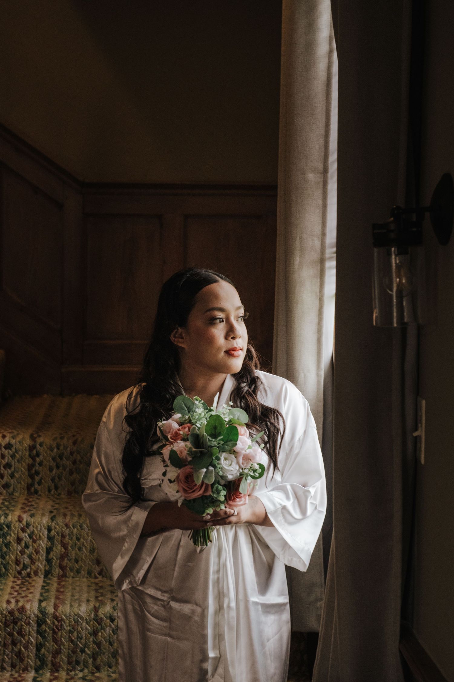A bride in a white robe holds a bouquet while gazing out a window in soft natural light.