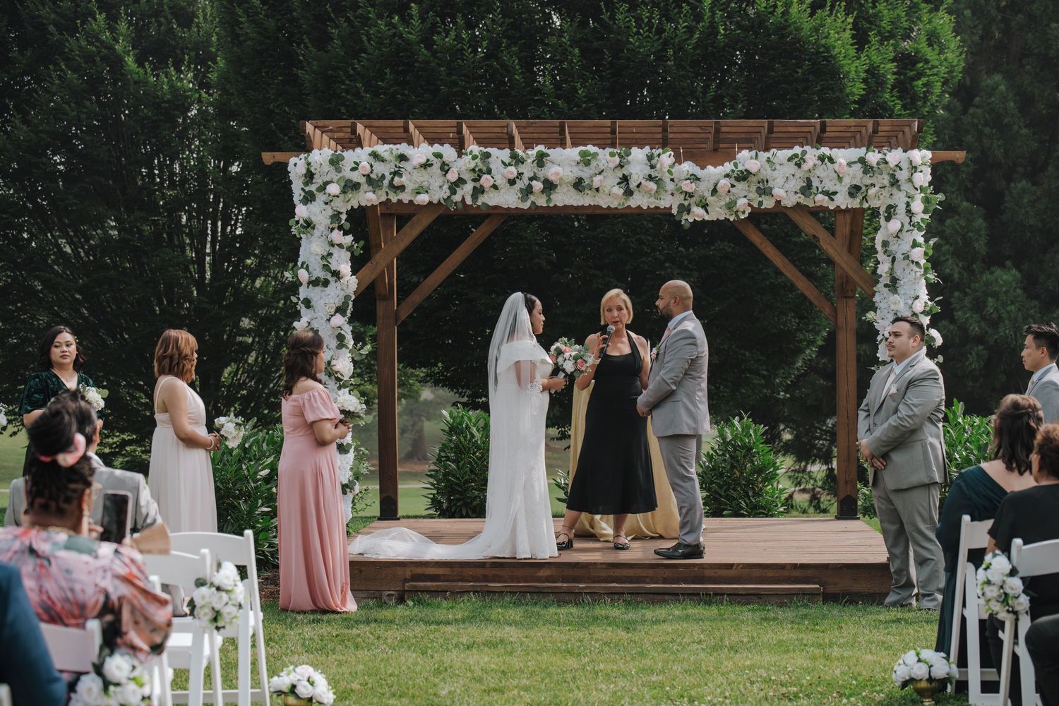 Wedding ceremony takes place under a rustic wooden pergola adorned with white flowers in an outdoor garden setting.
