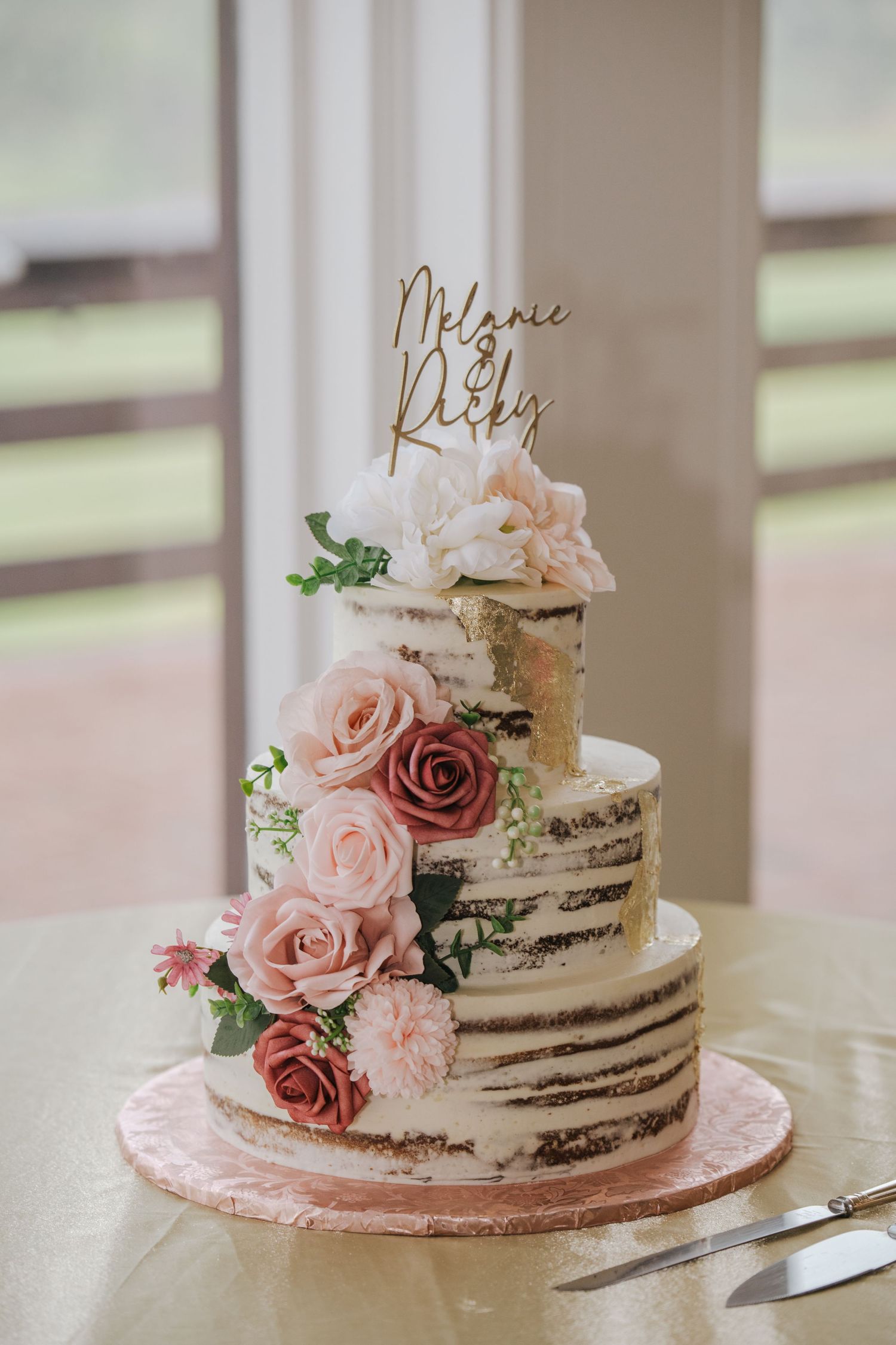 Semi-naked wedding cake decorated with pink roses and custom gold topper on display.